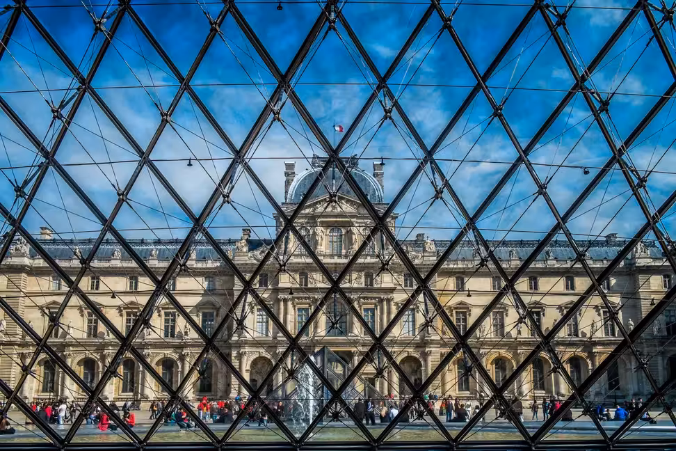 The Louvre palace seen through the glass and steel frame of the Pyramid, with visitors in the courtyard