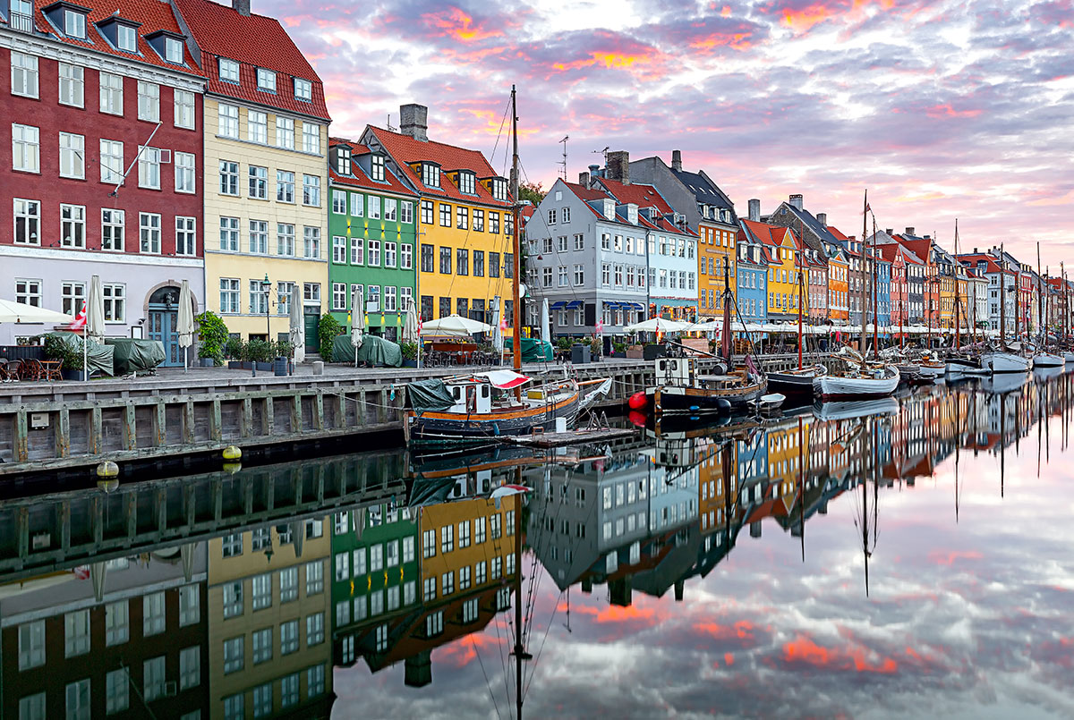 Copenhagen's Nyhavn Canal at sunrise features colorful historic houses lining the waterfront, reflecting their vibrant facades on the calm water, and a quiet city embankment, showcasing the iconic and picturesque Danish harbor.