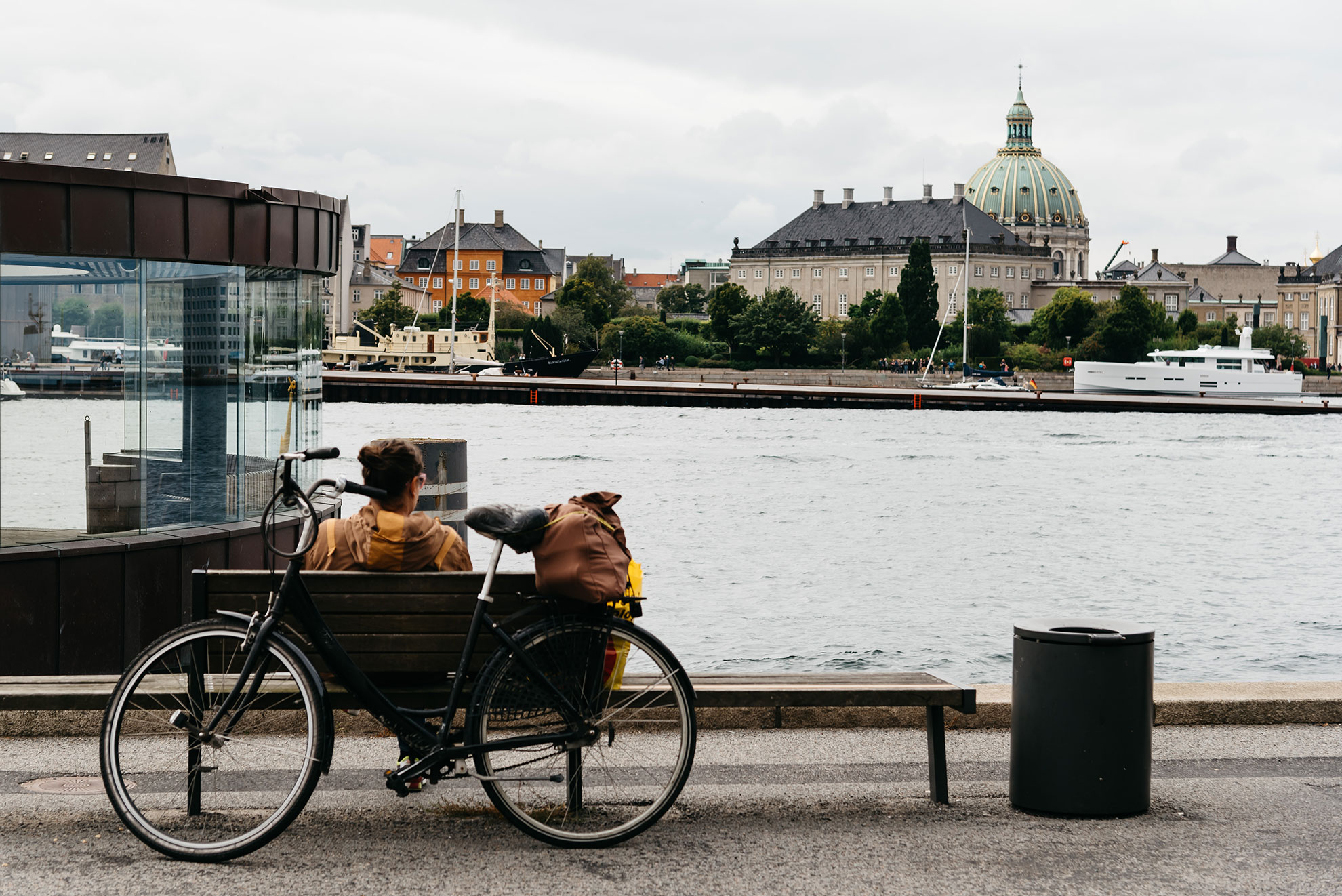 A woman traveler with a bicycle sits on a bench against the Copenhagen waterfront, enjoying the scenic view of the harbor and colorful buildings, embodying a relaxed, independent travel experience in Denmark.