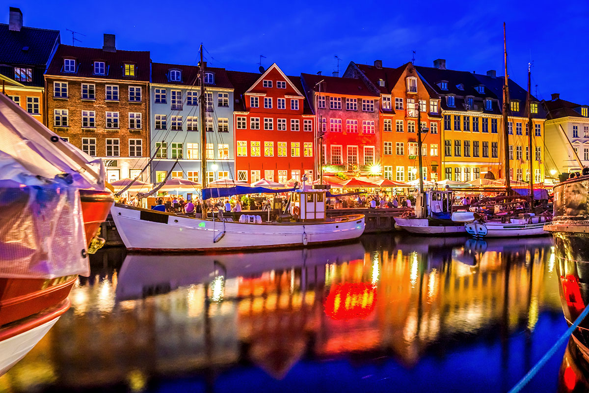 Denmark, Copenhagen, Nyhavn evening view features historic wooden boats docked along the canal, reflecting colorful facades of a row of houses illuminated by warm lights, creating a picturesque and vibrant harbor scene.