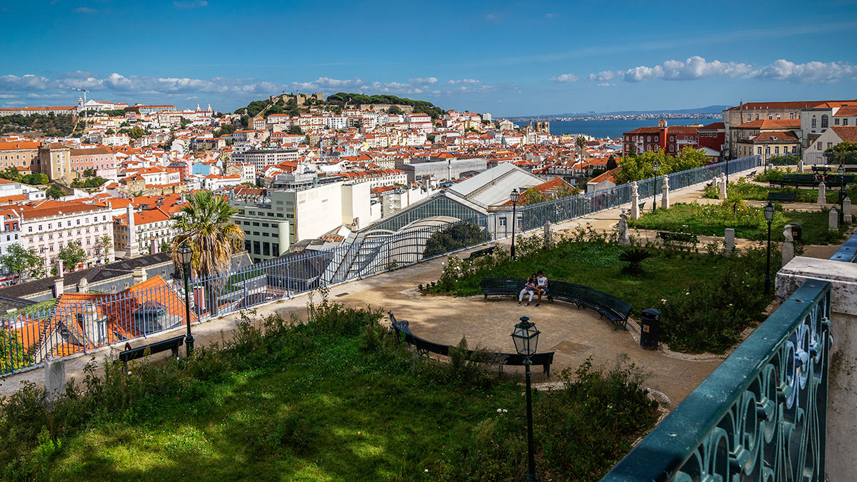 A picturesque scene from Miradouro de São Pedro de Alcântara in Lisbon, Portugal, features manicured gardens overlooking the city's sprawling landscape, with the São Jorge Castle visible in the distance.