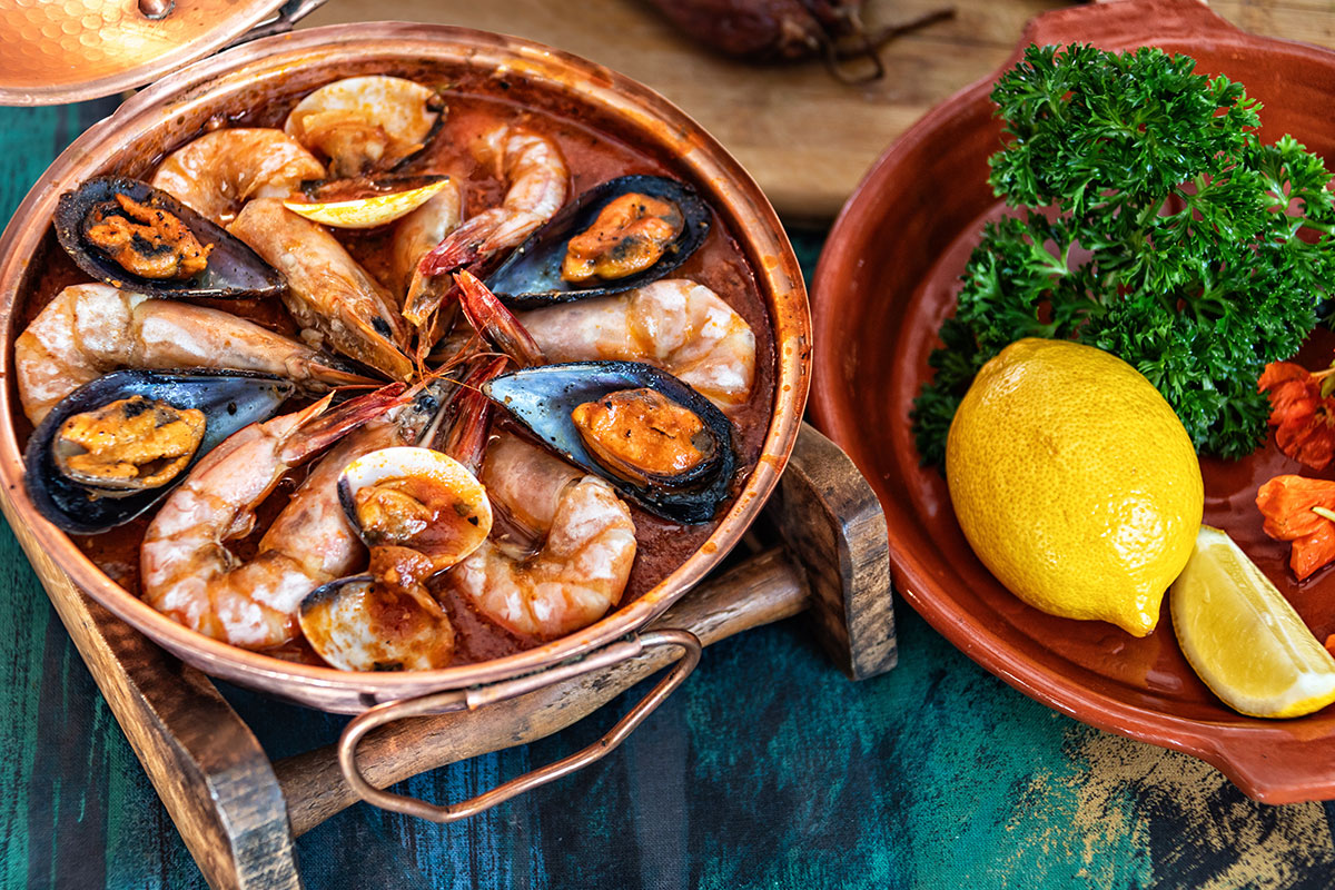 A steaming cataplana de marisco do Algarve, a traditional Portuguese seafood stew cooked in a copper clam-shaped pot, showcases a vibrant mix of shrimp, clams, mussels, and other shellfish in a flavorful broth, garnished with fresh herbs.