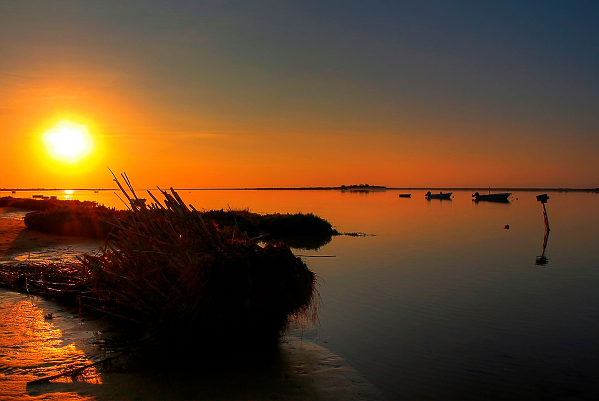Golden sunset over the calm waters of Ria Formosa, with silhouettes of boats and distant islands against a vibrant sky.