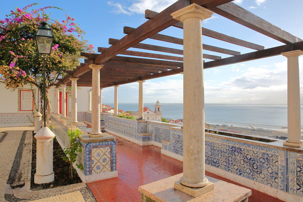 Scenic view from Miradouro de Santa Luzia overlooking Alfama's rooftops and the Tagus River, framed by azulejo tiles and blooming bougainvillea.