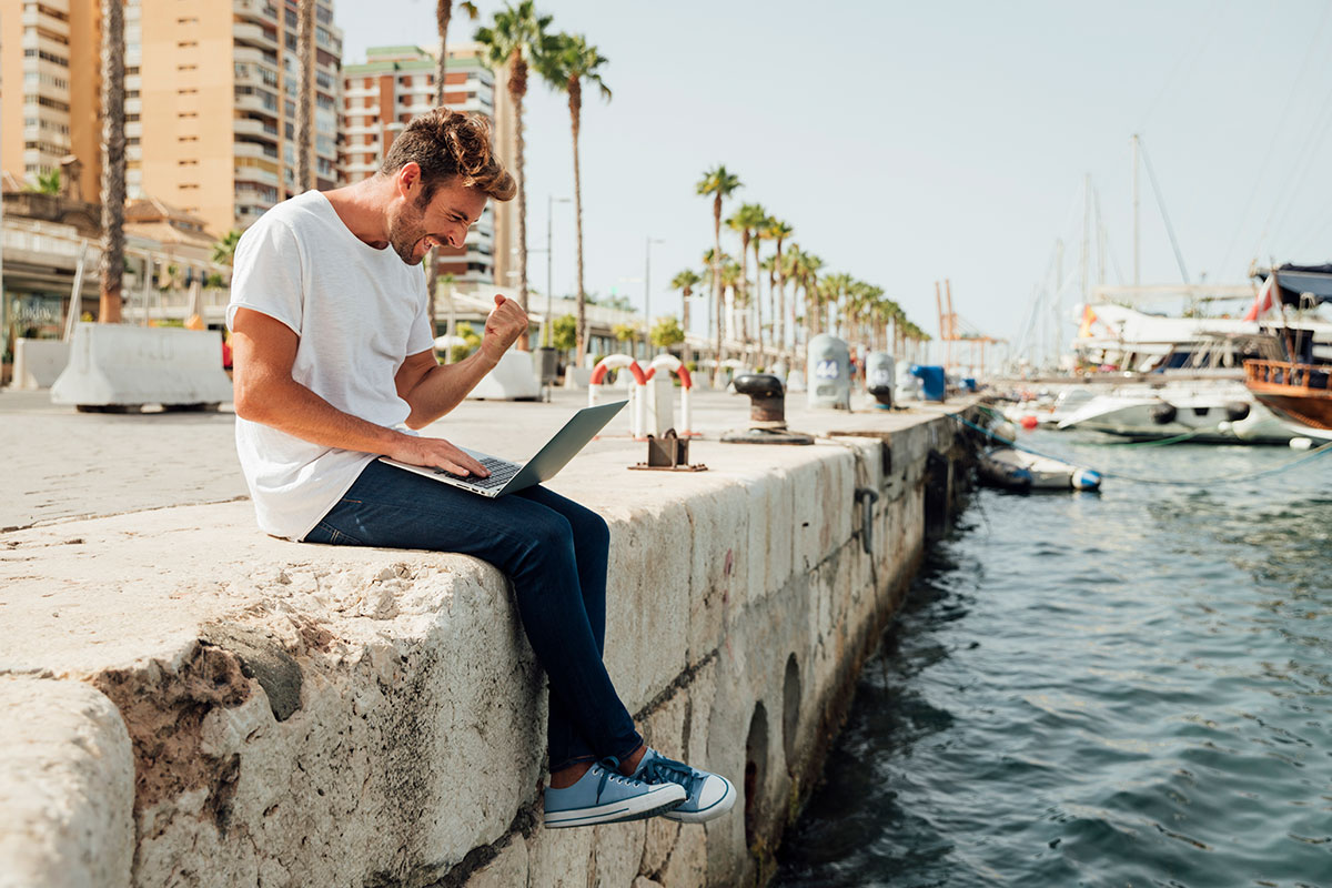Smiling man with a laptop raising his arms in celebration while sitting by the Tagus River in Lisbon, with cityscape and bridge in the background.