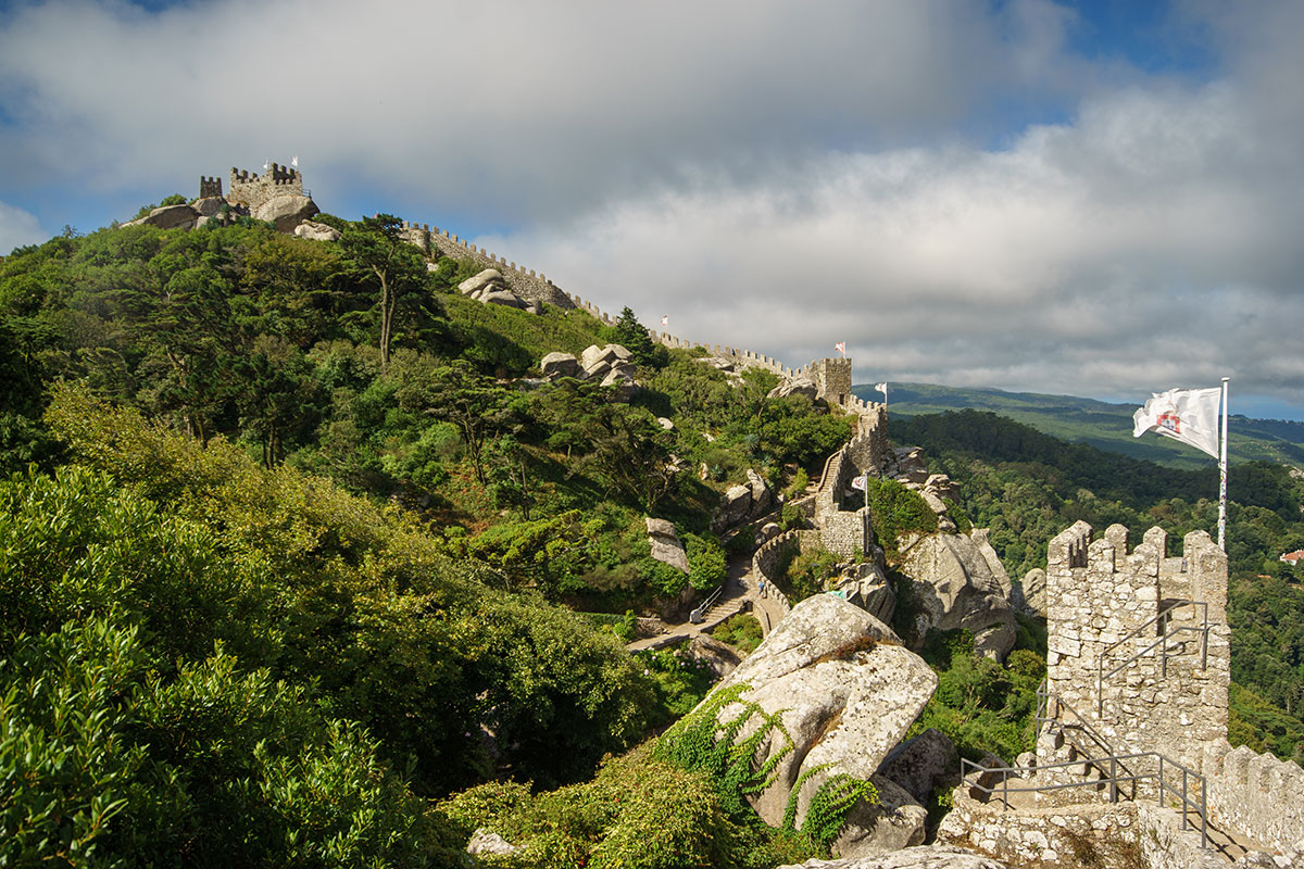 A panoramic view of the ancient Moorish Castle (Castelo dos Mouros) perched on a forested hilltop in Sintra, Portugal, with stone walls snaking along the ridge and views of the valley below.
