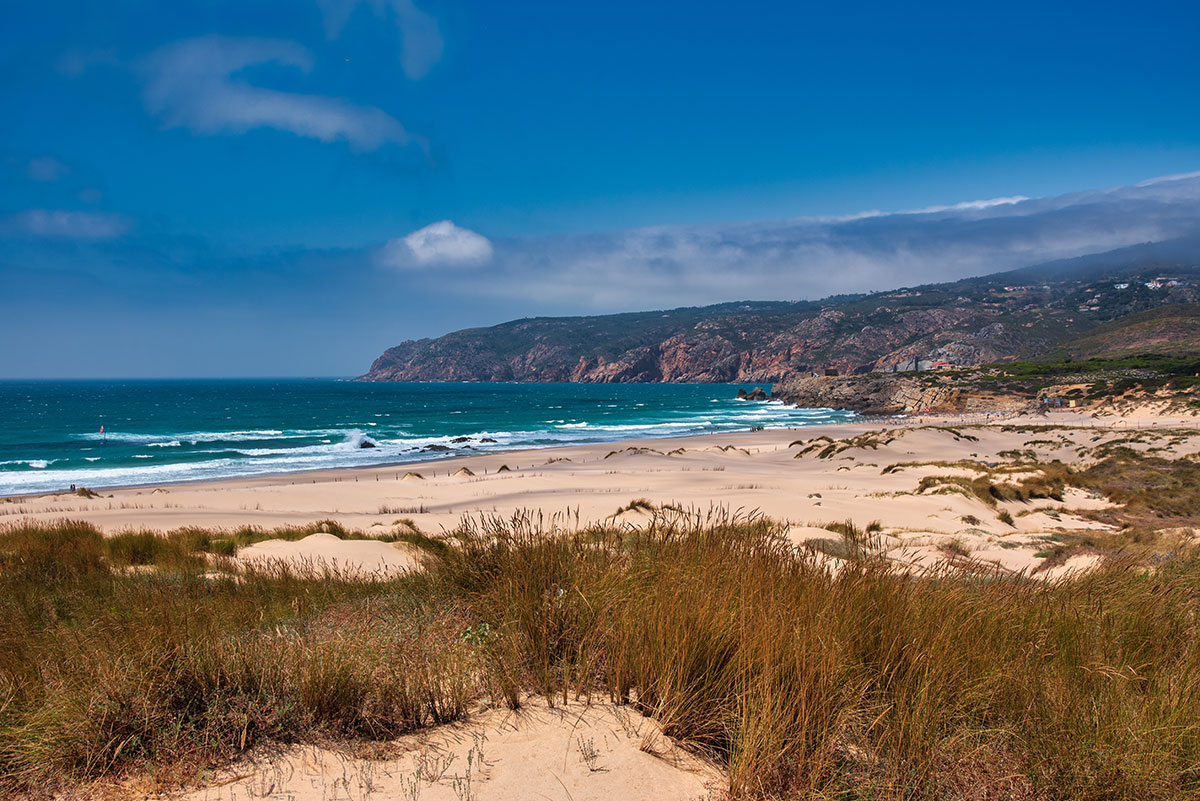 Wide view of Praia do Guincho near Cascais, Portugal, featuring golden sand dunes, crashing Atlantic waves, and rugged cliffs under a dramatic sky.