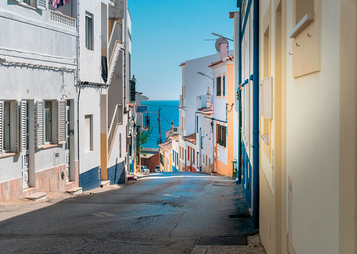Cobbled street in Burgau leading down to the ocean, with whitewashed houses and sea in the background.