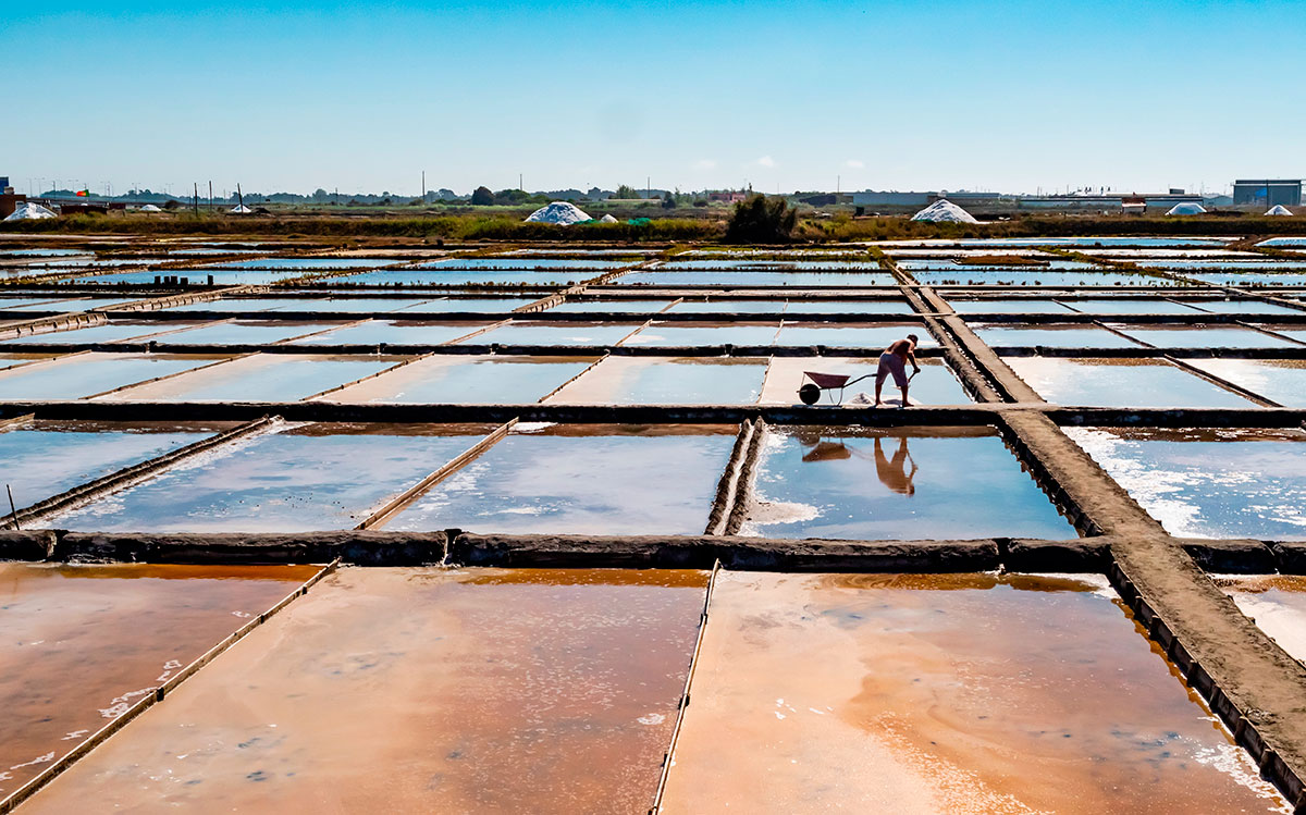 Colored salt ponds at a traditional salt extraction site in Aveiro, Portugal, with the silhouette of a worker harvesting salt at sunset.