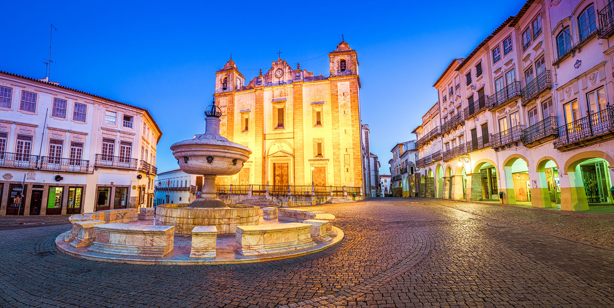 Praça do Giraldo at dusk with marble fountain and illuminated Igreja de Santo Antão, historic centre of Évora, Portugal