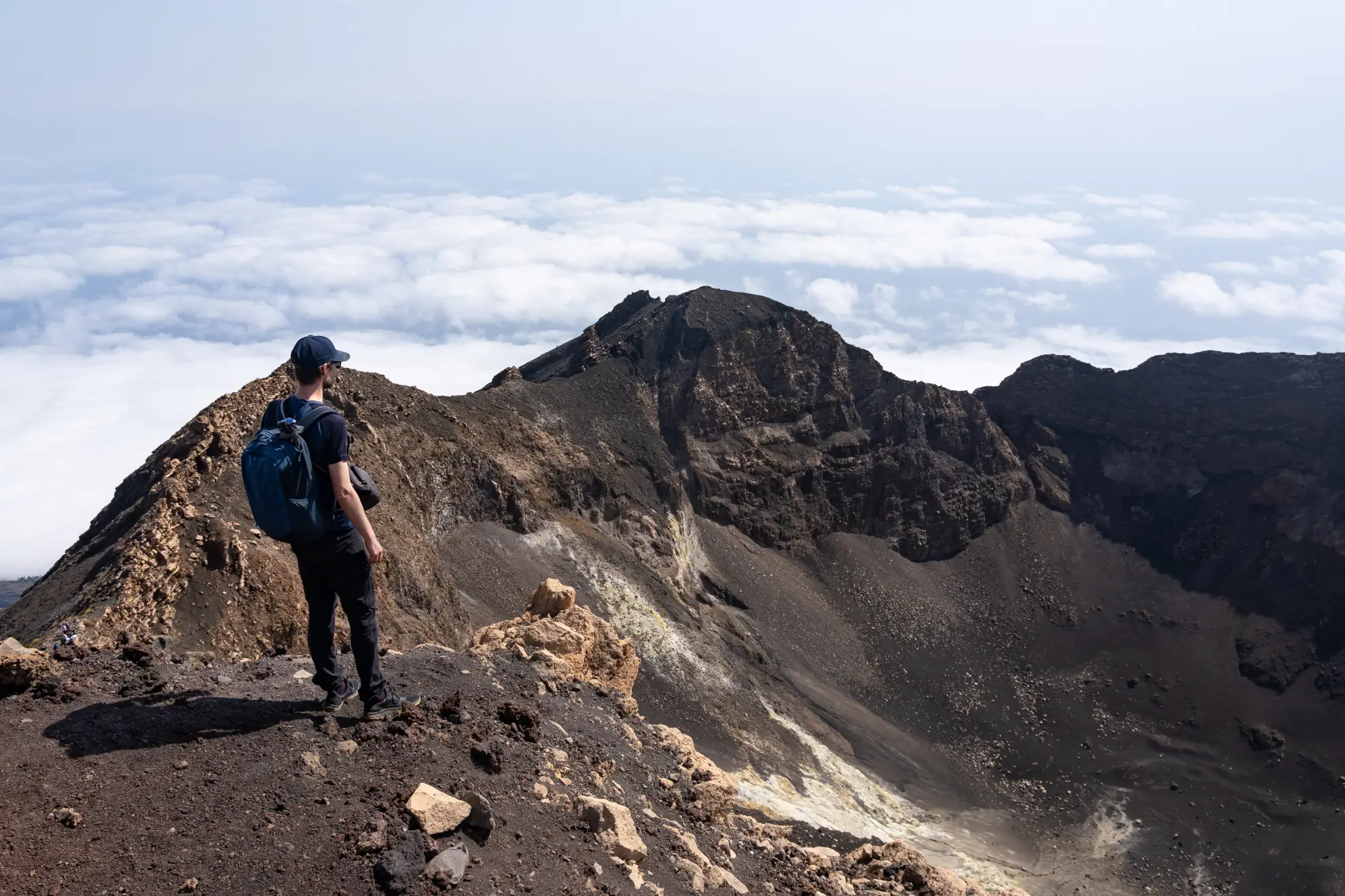 A hiker standing at the crater rim of Pico do Fogo volcano in Cape Verde, the vast caldera floor spread below with the smaller secondary cone visible and the Atlantic ocean in the distance