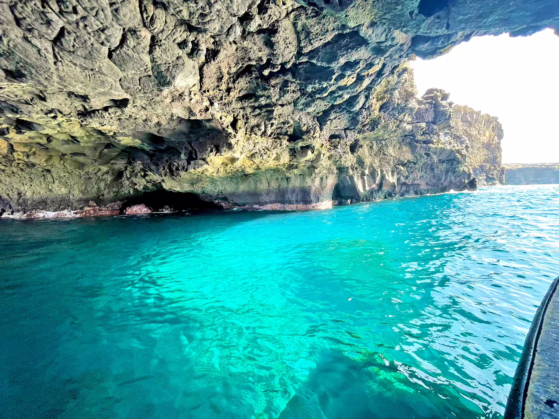 A small boat in a glowing turquoise sea cave on the Cape Verde coast, snorkellers visible in the clear water below the hull