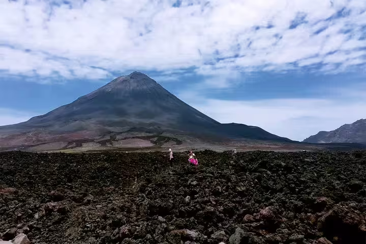 Hikers ascending the steep volcanic scree near the summit of Pico do Fogo, lava fields and the caldera floor far below them, the Atlantic visible on the horizon