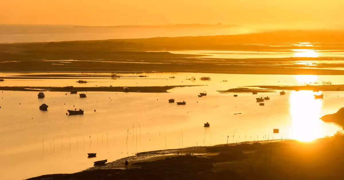 Wooden boat crossing Ria Formosa lagoon with Faro skyline and barrier islands visible.