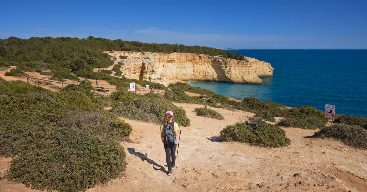 Hiker on sun-drenched Seven Hanging Valleys cliff path above turquoise sea, Algarve Portugal.