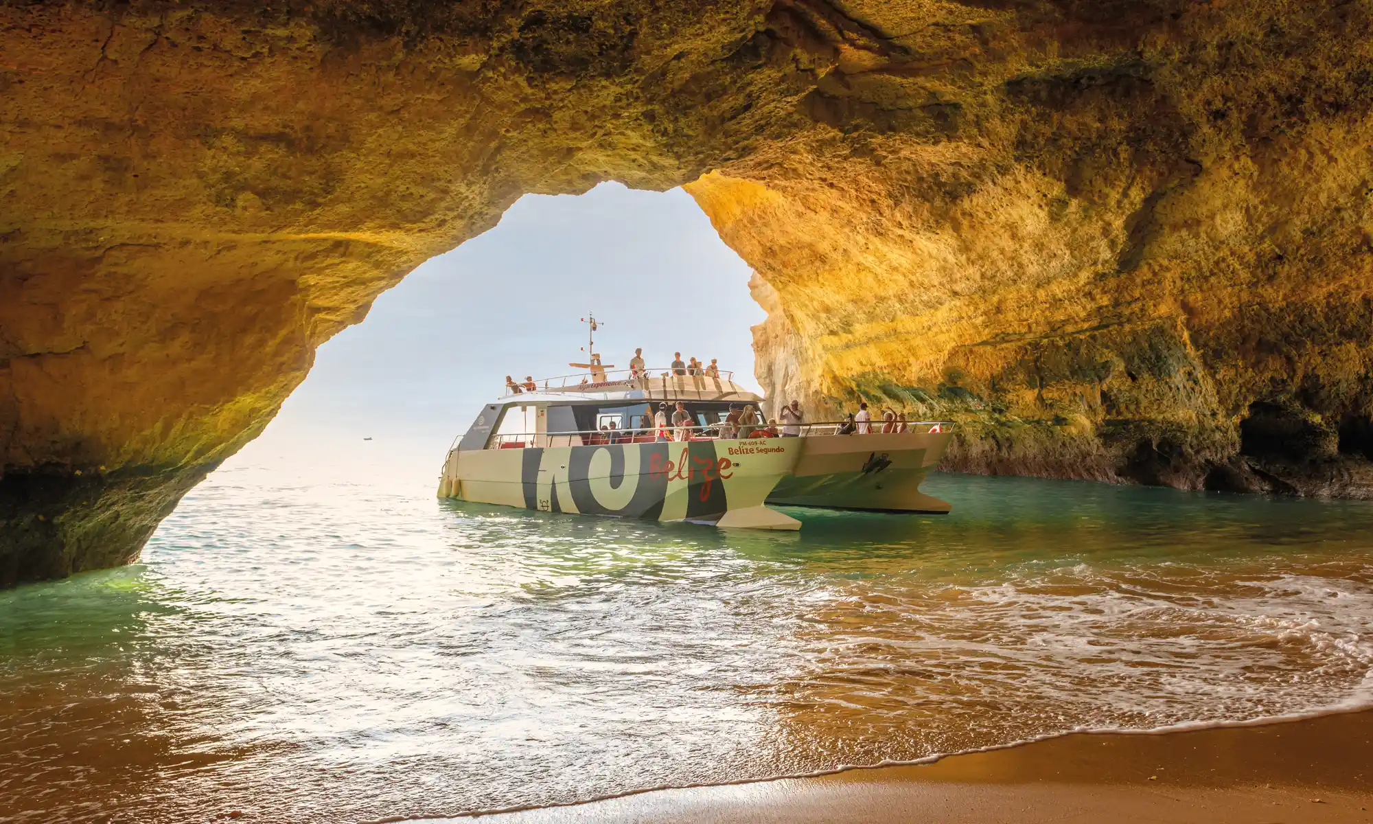 Catamaran approaching Algarve limestone cliffs with passengers on deck in sunlight.