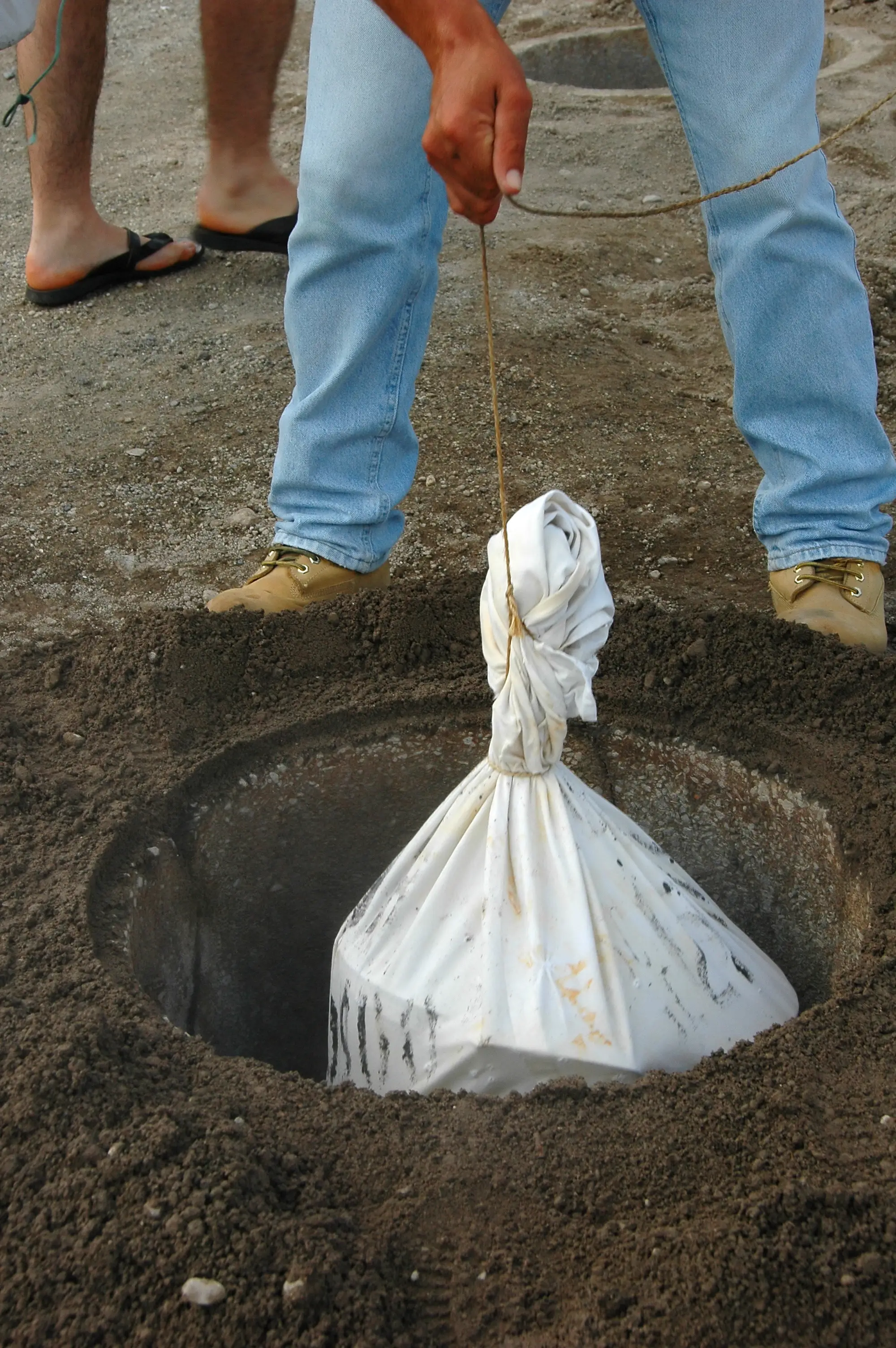 Worker lifting mud-covered burlap sack from geothermal pit at Furnas Lake during levantamento